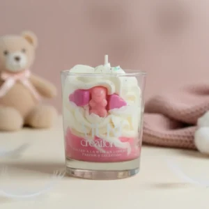 Bougie artisanale Amour de bébé de la marque Muri Créations aux notes douces et poudrées. Bougie gourmande en verre avec cire fouettée blanche, base rose et décorations ourson et cœurs, mise en scène avec un ours en peluche et des plumes pour une ambiance cocooning.