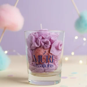 Bougie gourmande artisanale Muri Créations parfumée à la barbe à papa, composée de cire fouettée violette et d'un insert rose pailleté dans un verre transparent, sur fond pastel avec décoration festive.