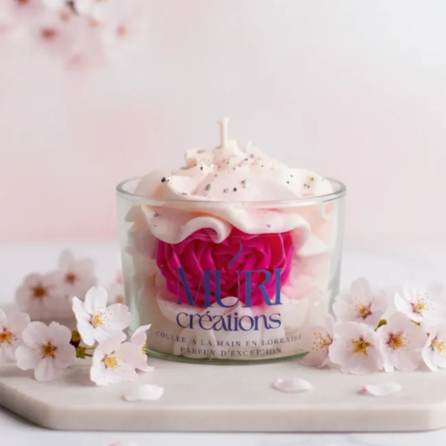 Bougie parfumée artisanale fleur de cerisier par Muri Créations, coulée à la main en Lorraine. Design gourmand en cire fouettée avec rose fuchsia incrustée, décorée de paillettes et entourée de fleurs de sakura sur fond rose.