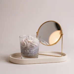 Bougie parfumée à l'ambre gris de la marque artisanale Muri Créations avec cire fouettée décorative, posée sur un plateau beige à côté d'un miroir doré pour une ambiance déco minimaliste.
