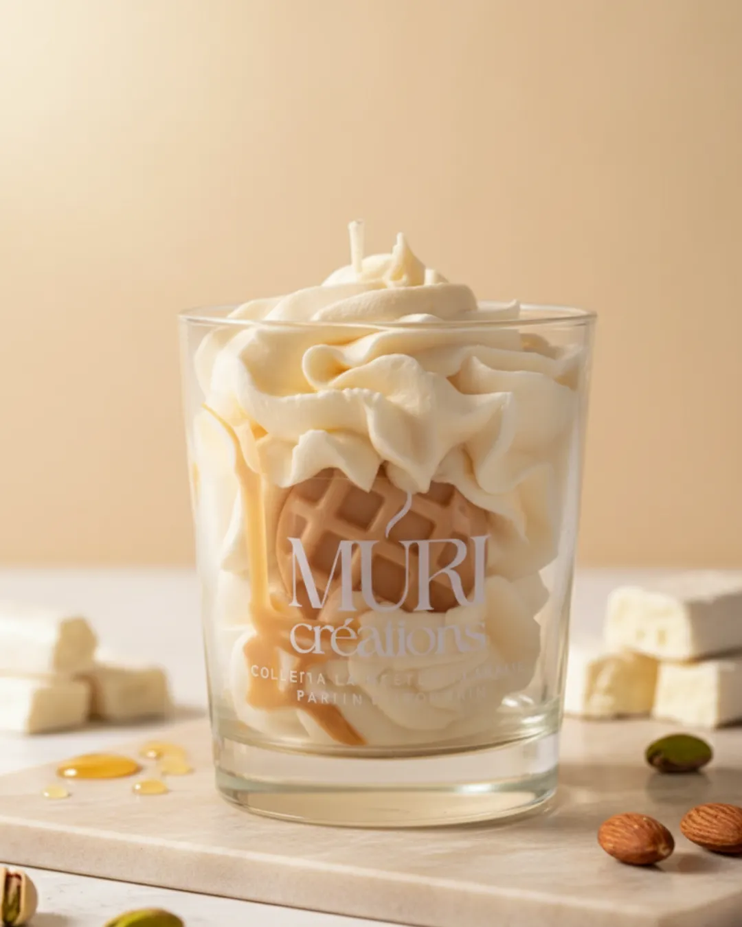 Bougie parfumée Nougat Blanc 1 Bougie parfumée au nougat de la marque artisanale Muri Créations, présentée dans un verre avec une cire effet chantilly, un insert gaufrette et un coulis caramel, entourée de morceaux de nougat blanc, d'amandes et de pistaches.