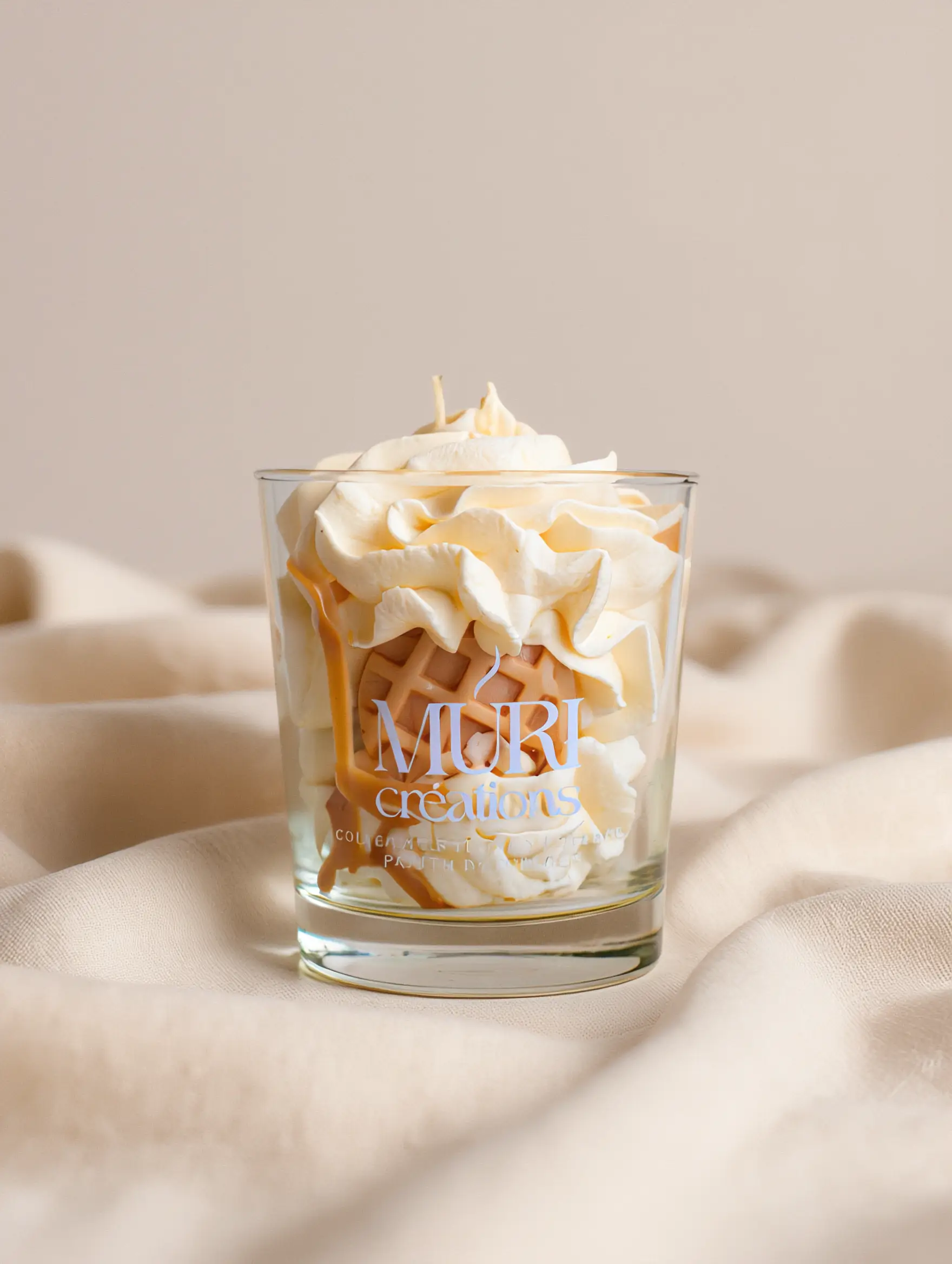 Bougie parfumée Nougat Blanc 4 Bougie parfumée au nougat de la marque artisanale Muri Créations, style gourmand avec cire fouettée façon chantilly, décoration gaufrette et coulis caramel dans un verre transparent sur fond beige.